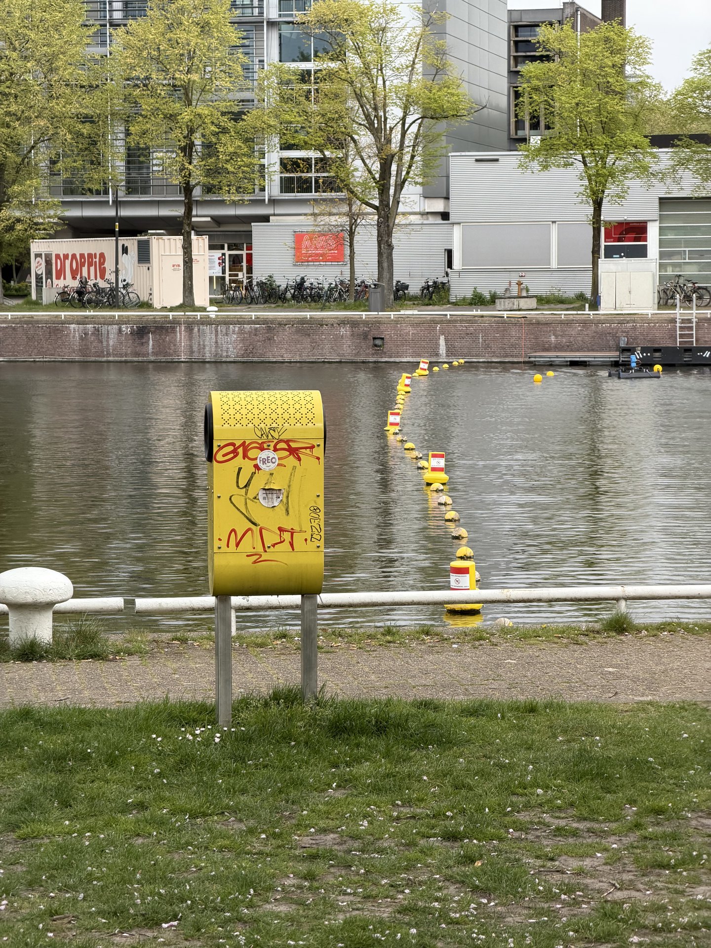 This photo captures a tranquil urban scene at Kade West, Marineterrein, Amsterdam, featuring a canal lined with modern buildings and vibrant green trees
